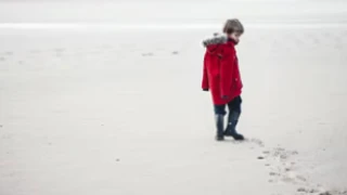 Little boy walking in a red jacket on the sand and turning back and looking at his footsteps